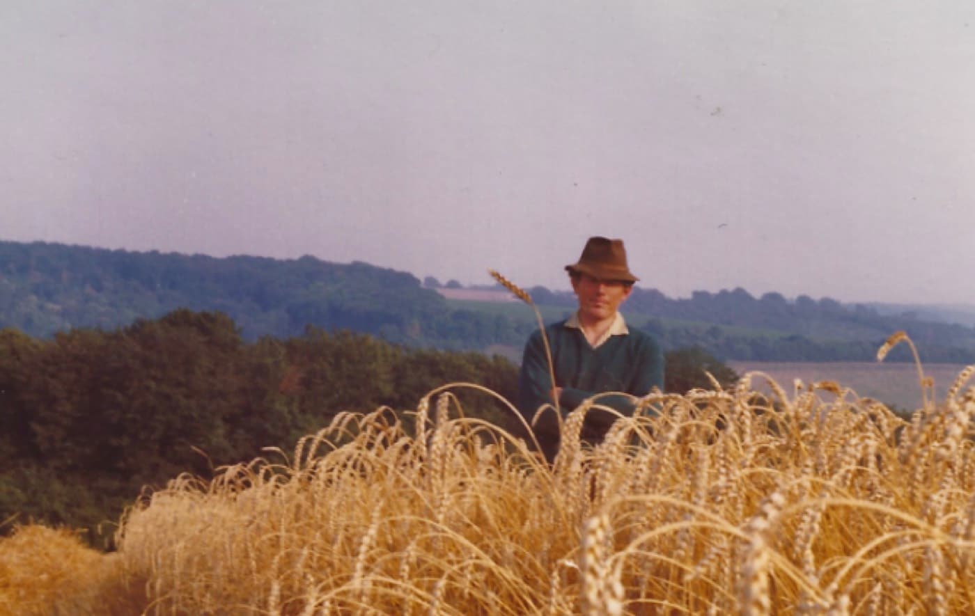 John Howard-Jones standing in a field of ripe wheat, wearing a hat, with wooded hills behind him.
